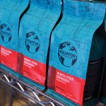 Photograph of Equator Coffee North Star Espresso beans lined up on a metal shelf. The bags are an indigo blue with bright read label with white writing.